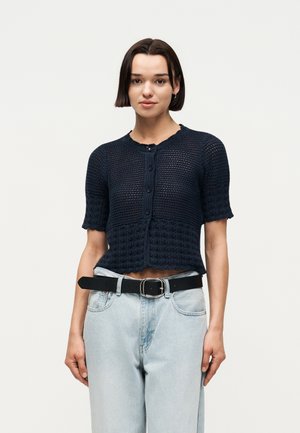 Jonge vrouw met kort donker haar die een marineblauw gebreid kort mouw vest draagt en lichtblauwe high-waisted jeans met een zwarte riem.