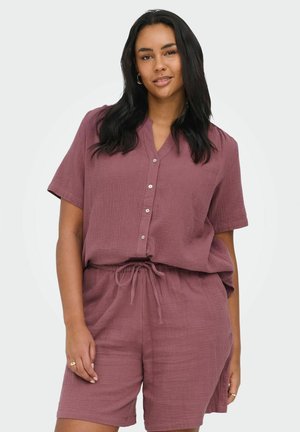 Naine, kellel on pikad tumedad juuksed, kannab lühikeste varrukatega nuppudega mauve-särki ja sobivaid nööriga lühikesi pükse, seisab valgel taustal.