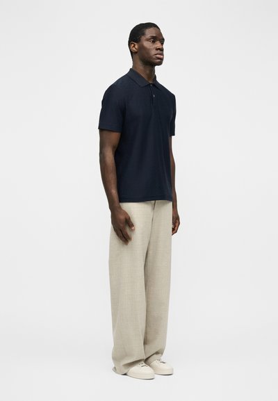 Άνδρας που στέκεται φορώντας μπλε ναυτικό πόλο με κοντά μανίκια, μπεζ παντελόνια φαρδιάς γραμμής και λευκά αθλητικά παπούτσια, σε απλό ανοιχτό φόντο.