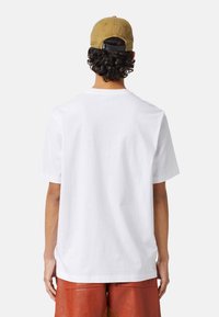 Biała bawełniana koszulka o luźnym kroju, z krótkim rękawem i okrągłym dekoltem, noszona z brązowymi skórzanymi spodniami i beżową czapką.