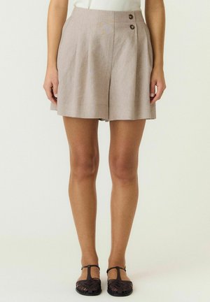 Beige shorts med høj talje med to sidelommer med knapper, båret med sorte vævede T-rem flade sko og en hvid top, ben synlige fra midt på låret og ned.