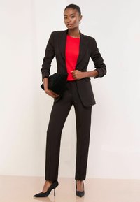 Fekete szabott öltöny, illeszkedő blézerrel és egyenes szárú nadrággal. Alatta élénkpiros felső, kezében egy fekete kesztyűtartó. Elegáns magassarkú cipők.