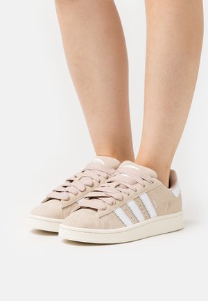 Beige suède sneakers met witte strepen en dikke zolen, gedragen aan de voeten, afgebeeld tegen een effen witte achtergrond.