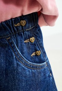 Pantalones de mezclilla con una cinturilla elástica que cuenta con tres botones en forma de corazón, de color azul oscuro, y costuras visibles en el bolsillo y la cinturilla.