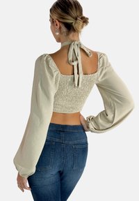 Top beige corto con espalda fruncida y mangas largas abullonadas; incluye un lazo en el cuello. Combinado con jeans de cintura alta azules.