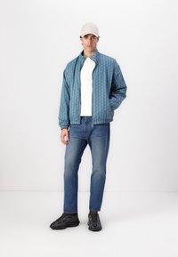 Teal patterned bomber jacket with ribbed cuffs and collar, worn over a white shirt, paired with blue jeans and black sneakers.