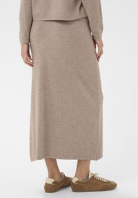 Persona vistiendo una falda midi de punto beige y un top a juego, combinados con zapatillas casuales marrones y crema, de pie sobre un fondo blanco.