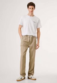 Beige drawstring pants with a relaxed fit, paired with a white crew neck t-shirt featuring a small logo. Sneakers with black accents.