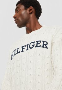 Cremfarbener, kabelgestrickter Pullover mit dunkelblauem, gesticktem "HILFIGER"-Logo auf der Brust. Runder Ausschnitt, strukturiertes Muster, lässige Passform.
