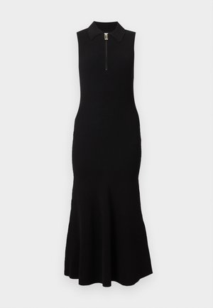 Moteris dėvi juodą rankovių midi suknelę su užtrauktuku priekyje ant apykaklės ir juodus aukštakulnius sandalų su raišteliais, stovinti prieš baltą foną.