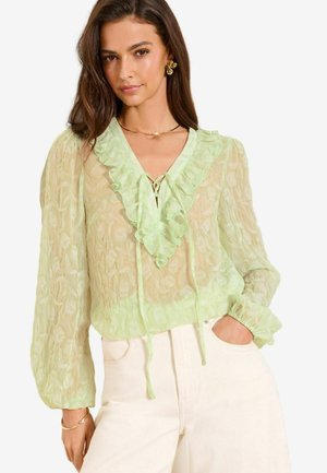 Žena oblečená v priesvitnej svetlozelenej blúzke s volánovým výstrihom do V a dlhými nadýchanými rukávmi, vstrčená do vysokých bielych nohavíc, stojaca s jednou rukou v vrecku.