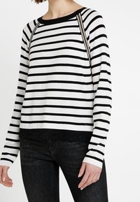 Chemise à manches longues rayée avec des rayures horizontales noires et blanches, un col rond et une touche de dentelle noire au niveau de l'ourlet.