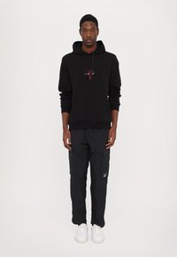 Sweat à capuche noir avec un design brodé rouge, associé à un pantalon cargo sombre avec des poches. Modèle portant des baskets blanches, se tenant devant un fond uni.