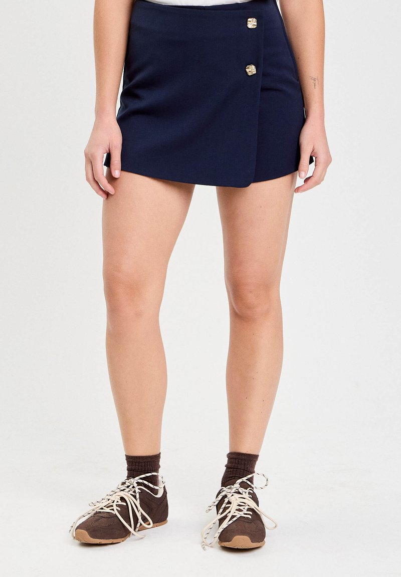 Kobieta w granatowej spódnicy z zakładką z złotymi guzikami, brązowych skarpetkach i brązowych tenisówkach z białymi sznurówkami na białym tle.