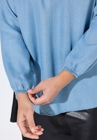 Camicia di jeans azzurro chiaro, comoda e morbida, con polsini elasticizzati, cuciture visibili e orlo inferiore arrotondato. Indossata su una gonna nera.