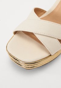 Beige canvas wedge sandal with crossed straps, a smooth square toe, and a layered gold-accented sole. Sturdy and textured surface.