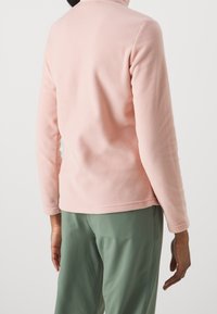 Ljusrosa fleecejacka med hög krage, med långa ärmar och en slät textur, tillsammans med gröna byxor.