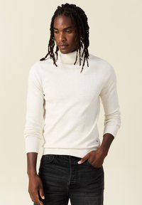 Suéter de cuello alto beige claro en punto suave, diseño ajustado, con puños y dobladillo acanalados, combinado con jeans de mezclilla oscura.