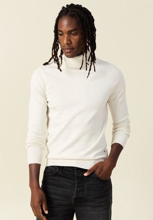 Un pull roulé beige clair en tricot doux, avec une coupe ajustée, des poignets et un ourlet côtelés, associé à un jean en denim foncé.