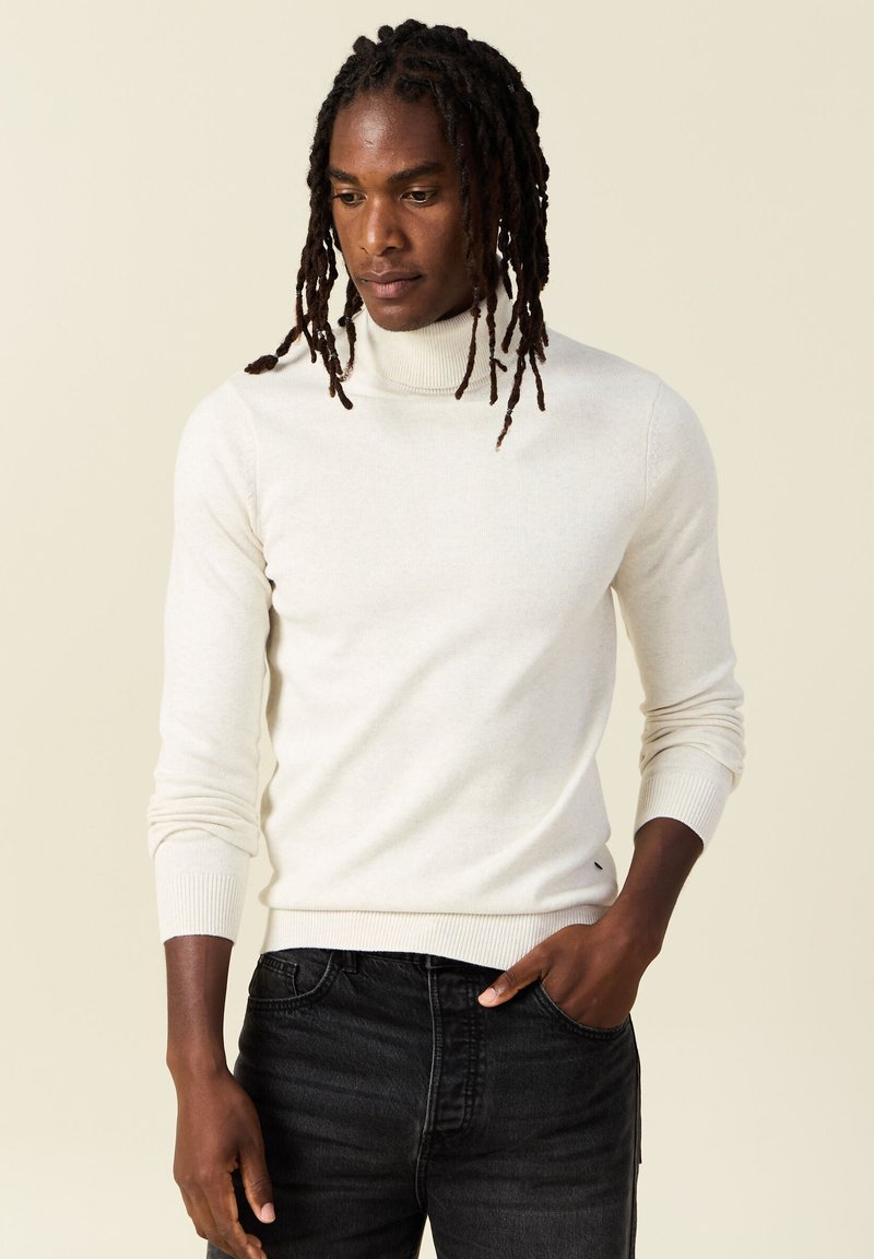 Suéter de cuello alto beige claro en punto suave, diseño ajustado, con puños y dobladillo acanalados, combinado con jeans de mezclilla oscura.