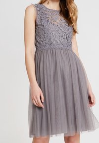 Robe grise avec un corsage en dentelle à motifs floraux, sans manches, et une jupe en tulle à plusieurs couches. Texture douce et longueur au genou.