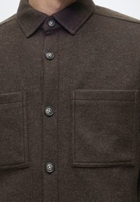 Gros plan d'une chemise marron foncé à boutons, avec deux grandes poches poitrine et un col classique.