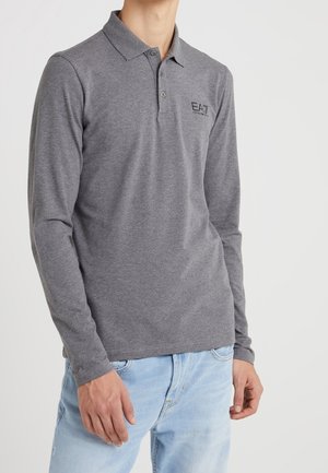 Homme portant un polo gris à manches longues avec le logo EA7 et un jean bleu clair, debout devant un fond blanc uni.