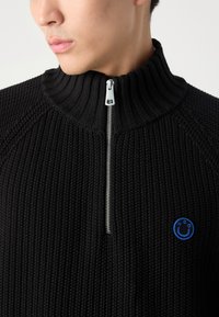 Maglione nero a zip con colletto a coste, caratterizzato da una cerniera argentata e un logo circolare blu sul davanti. Tessuto texturizzato.