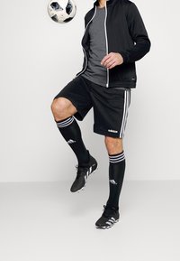 Schwarze Sportjacke mit weißem Reißverschluss, graues T-Shirt, schwarze Shorts mit weißen Streifen, schwarze Socken und schwarze Fußballschuhe, mit einem Fußball.