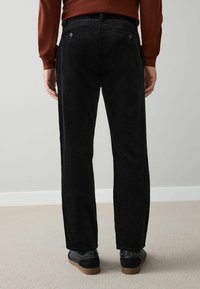 Black corduroy pants with wide legs, featuring two back pockets with blue buttons and a textured surface, paired with black shoes.