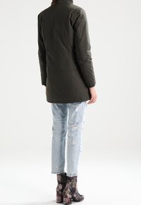 Parka vert olive à coupe droite, associée à un jean bleu clair déchiré et des bottines à talons fleuries. Col au design simple.