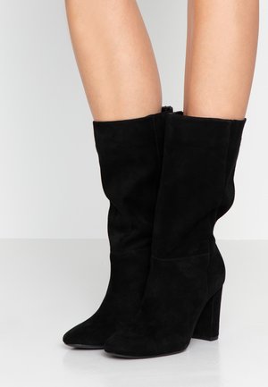 Vue des jambes d'une personne portant des bottes en daim noir mi-mollet à talons carrés, debout sur une surface blanche lisse et réfléchissante.