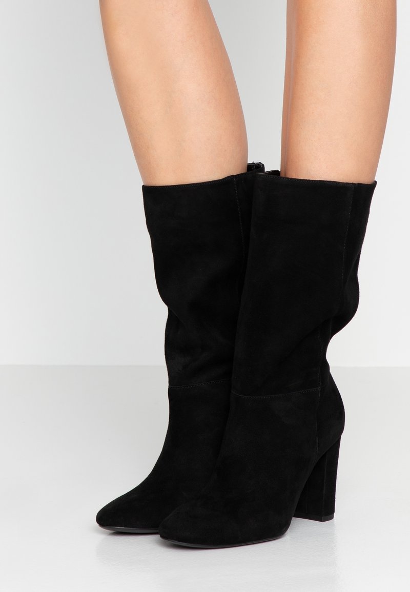 Vue des jambes d'une personne portant des bottes en daim noir mi-mollet à talons carrés, debout sur une surface blanche lisse et réfléchissante.