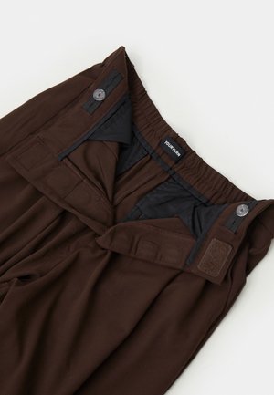 Bruine broek met een elastische tailleband, verstelbare knoop en klittenbandsluitingen, en een zichtbaar merkembleem binnenin de tailleband.