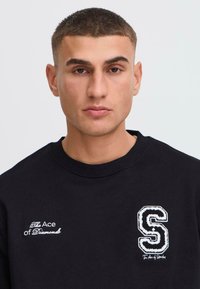 Hanorac negru cu texte și modele brodate albe, guler rotund și o textură moale, cu logo-uri pe partea stângă a pieptului.