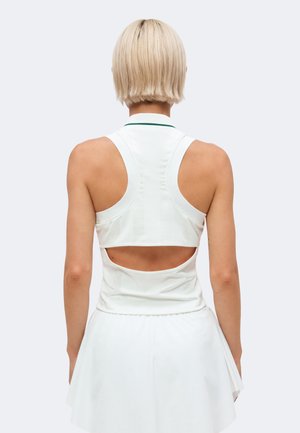 Femme aux cheveux blonds courts portant un haut blanc sans manches avec une découpe dans le dos et une jupe plissée blanche, dos à l'appareil photo.