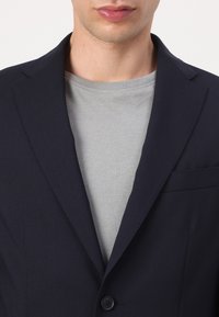 Sötétkék zakó, slim fit szabással, texturált anyagból, bemetszett gallérral, világosszürke kerek nyakú póló felett. Elöl zsebe és gombos záródása van.