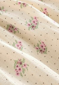 Baumwollstoff mit kleinen braunen Punkten auf einem cremefarbenen Hintergrund, ergänzt durch verstreute rosa Blumenmuster und grüne Akzente.