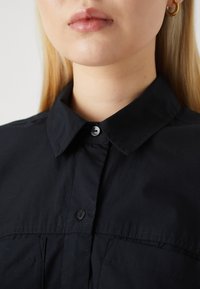 Juodas marškiniai su aštriu apykakle, pasižymintys glotnia tekstūra ir subtiliu siuvinėjimu. Įtraukti mygtukai ir priekinės kišenės detalės.