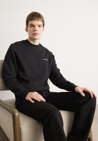 Crna dukserica s okruglim izrezom i dugim rukavima, s malim bijelim Calvin Klein logotipom na lijevoj strani prsa. Nošena s crnim hlačama.