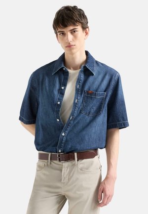 Junger Mann trägt ein aufgeknöpftes kurzärmeliges Jeanshemd über einem weißen T-Shirt, beige Hose und braunen Gürtel, steht vor schlichtem Hintergrund.