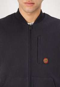 Black bomber jacket with a high collar, zip closure, textured fabric, and a red and yellow "Santa Cruz" logo patch on the left side.