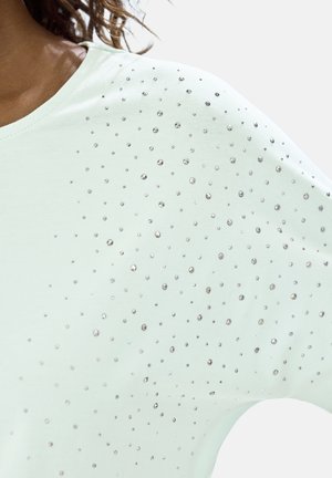 Close-up of a pale mint green top with scattered small silver rhinestones on the shoulder and upper sleeve of a person with dark curly hair.