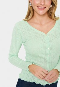 Cardigan long en vert clair, fabriqué en tissu texturé, avec un col en V, fermeture à boutons, et un ourlet et des poignets à volants.