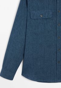 Camicia in denim blu a maniche lunghe con taschino frontale e chiusura con bottoni su uno sfondo bianco.