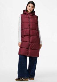 Mujer vestida con un chaleco acolchado largo burdeos con capucha sobre una camiseta blanca de manga larga, pantalones vaqueros anchos azules y zapatos casuales marrones, de pie de manera relajada.