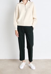 Sweter zapinany na zamek w kolorze kremowym, żakardowe spodnie w ciemnozielonym kolorze i białe trampki z wiązaniem, stojące na neutralnej podłodze.