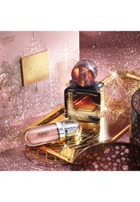 Flacon de parfum Kiko Milano Oud Escape et gloss à lèvres rose sur un plateau doré avec un arrière-plan rose pailleté et un emballage décoratif derrière.