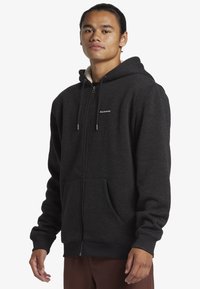 Felpa nera con zip, dall'interno morbido, cappuccio con coulisse, tasche anteriori e polsini a costine. Presenta un piccolo logo sul petto.