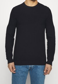 Mörkblå stickad tröja med långa ärmar, rundad halsringning, texturerad tyg och ribbade manschetter och nederkant. Bärs över ljusblå jeans.